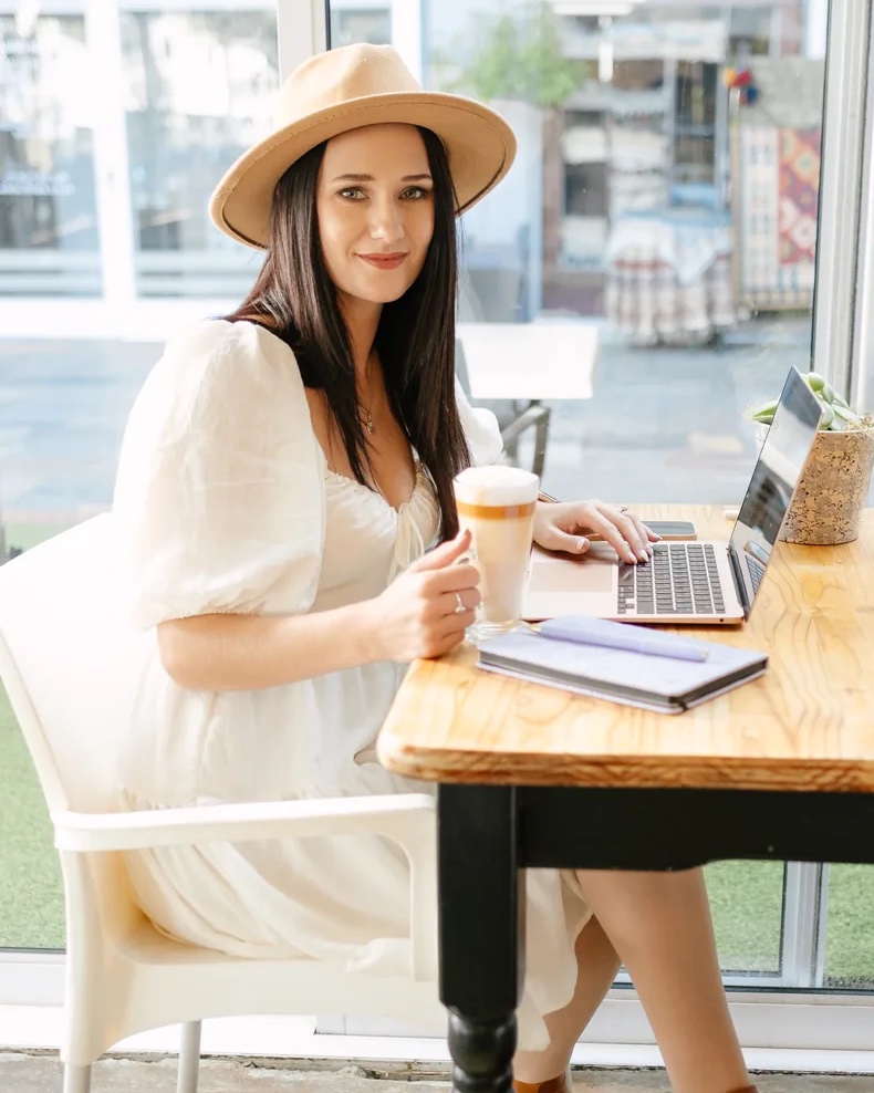 Liezle Potgieter at a cafe table with a laptop and a coffee, smiling at the camera.